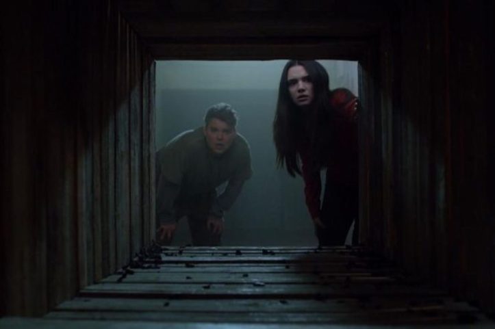 Image: A young woman with pale skin and long dark hair and a young man with light skin and blonde hair face the viewer as they peer down a dark tunnel.