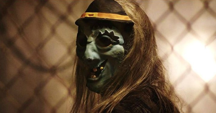 Image: A person with long blonde hair and a green witch mask looks over their shoulder at the viewer with a chain-link fence in the background.
