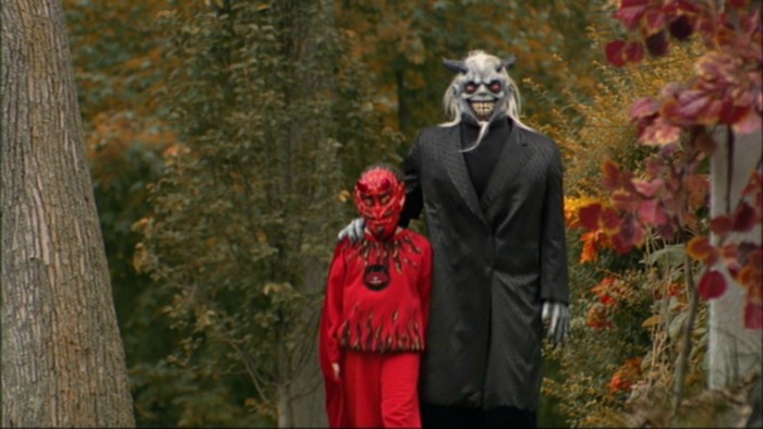 Image: A little boy in a red devil costume and a man in a pale demon mask and black coat walk down a street with trees and foliage surrounding them.