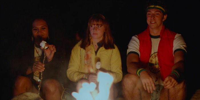 Three campers sit in front of a campfire. A Black woman in a jacket puts a marshmallow on a stick while a white woman with a yellow shirt and a white man with a red vest look on.