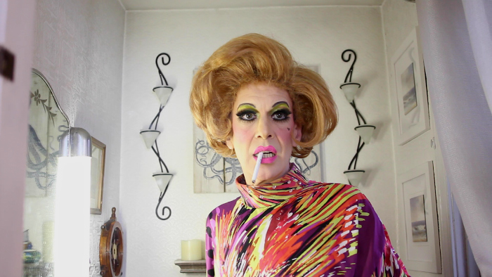 Image: A drag queen wearing a brightly colored caftan stares off camera with a cigarette dangling out of her mouth. She sits in front of a white wall with spare white decorations.