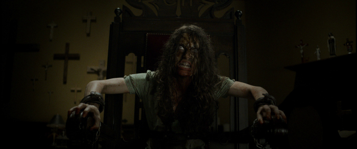 Image: A woman with a dirty, bloody face sneers off camera. She sits in a large chair with her wrists tied to the arms and crosses cover the wall behind her.