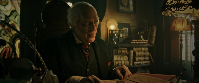 Image: A man in a three-piece suit sits at a desk with Art Deco lamps and a feathered quill. He is surrounded by books and stained glass windows.