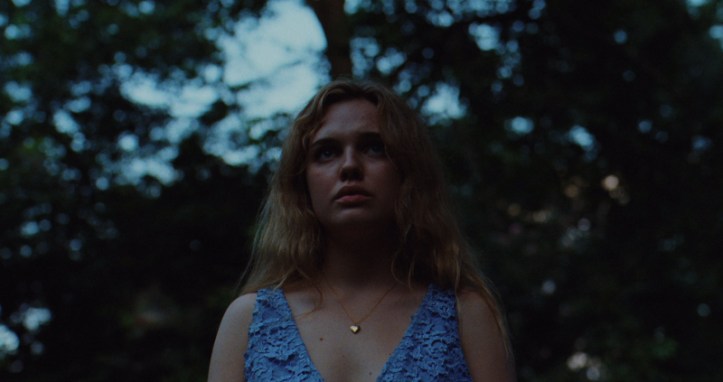Image: A teen girl with light skin, blonde hair, and a blue dress stands in a forest. She faces the camera and looks toward the horizon.