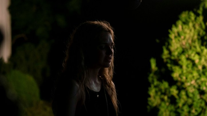 Image: A teenage girl with long blonde hair stands in front of some trees at night. Her face is mostly in shadow.
