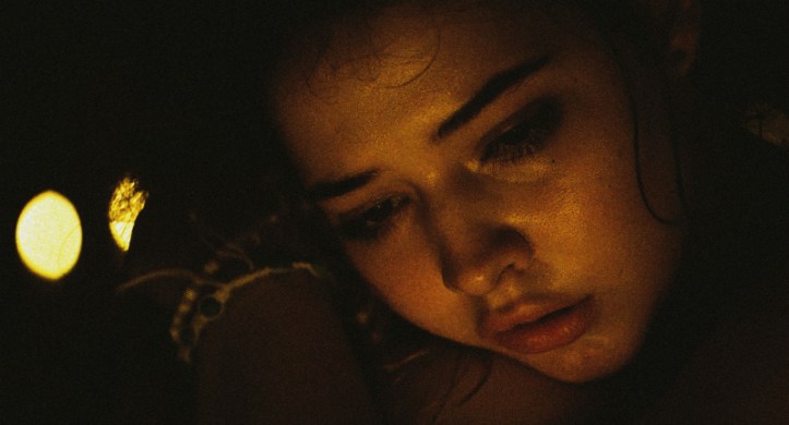 Image: A teen girl with light skin and dark hair sits with her head resting on her hands. Two lights shine behind her.
