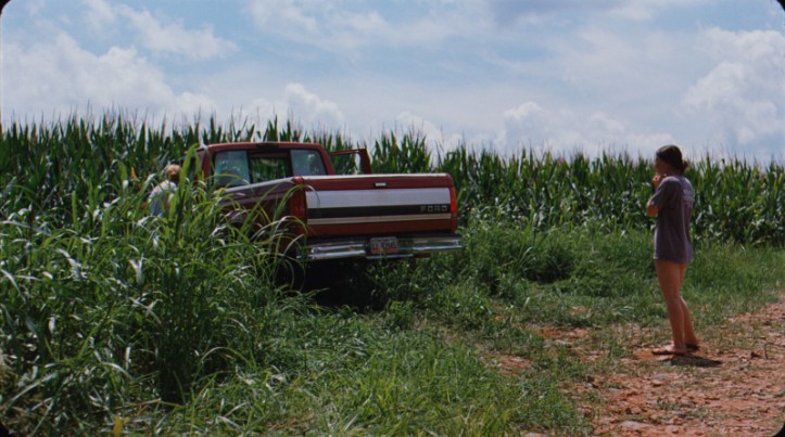 Image: A red pickup truck sits in a field with its doors open. A blonde girl peers inside the cab while a brunette girl stands behind the truck, looking worried.