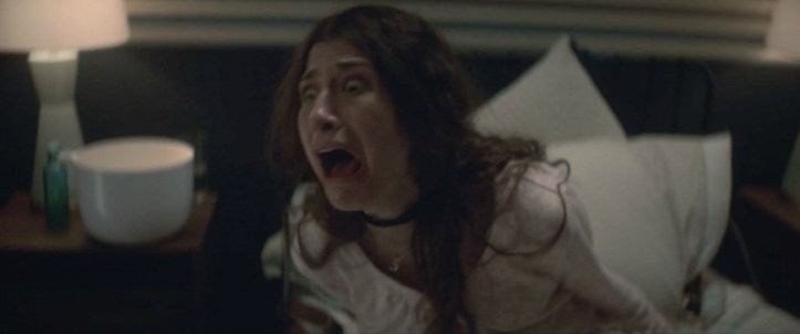 Image: A woman with long dark hair sits up in a bed and screams in pain and terror.