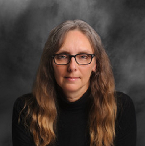 Image: A white woman with brown hair, black glasses, and a black sweater faces the camera in front of a grey and white backdrop.