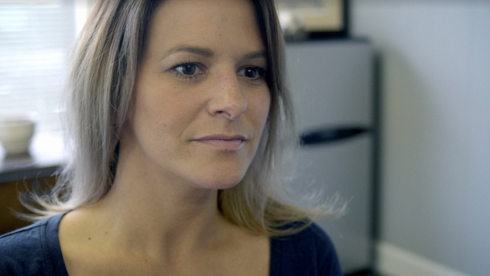 A woman with shoulder-length hair and a blue shirt sits in an office and looks at someone off-camera.
