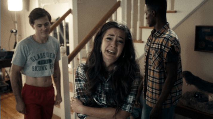 A still from Death Cast. A girl stands in a room with her arms crossed and a tearful, frightened look on her face. Two boys stand behind her looking away. 