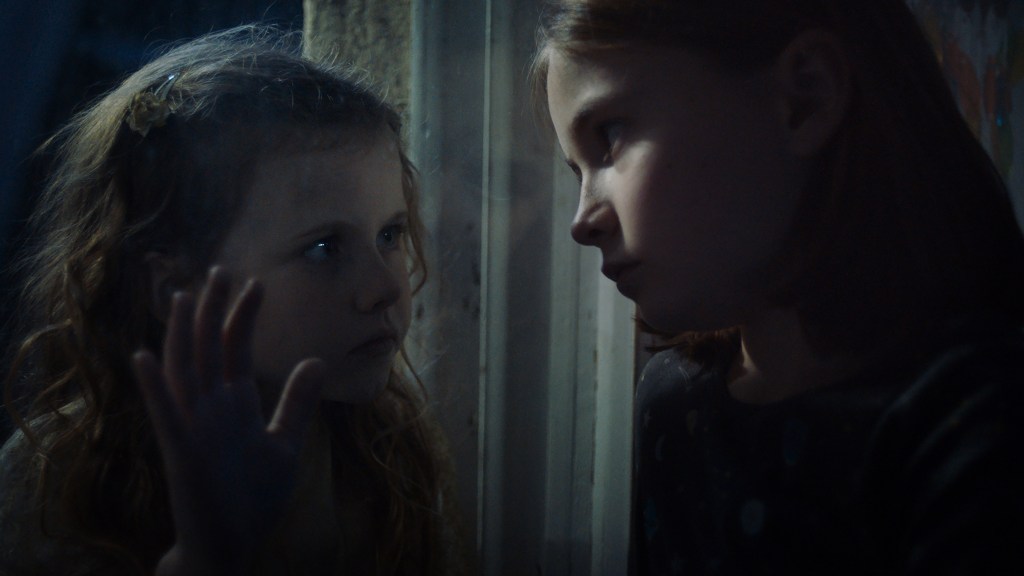 A still from Martyrs Lane. Two young girls face each other from opposite sides of a window.