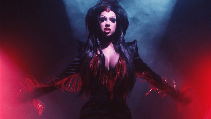 A still from So Vam. A person in a black jacket with red fringe stands with their arms outstretched in front of red and white lights against a black background.