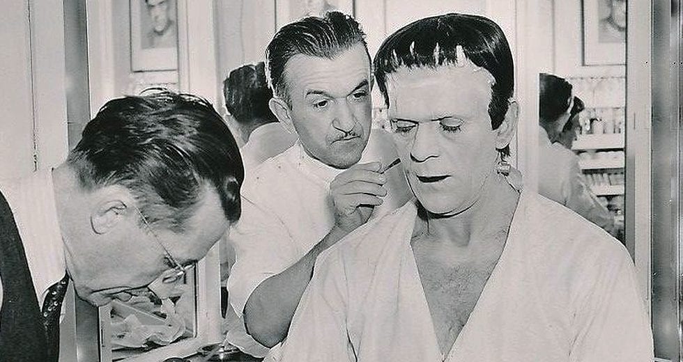 A behind-the-scenes photo of Jack Pierce applying Frankenstein makeup to Boris Karloff.