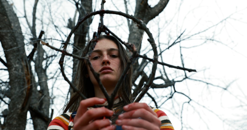 A still from Hellbender. Izzy stands in front of a leafless tree against a white sky. She holds a sigil made of twigs.