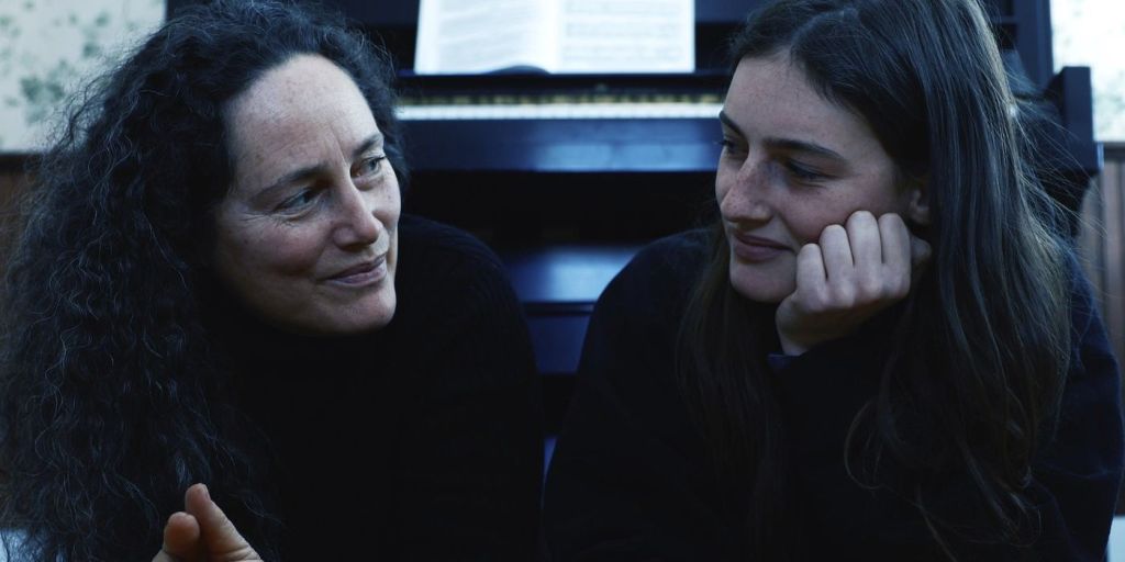 A still from Hellbender. Izzy and her mother sit in front of a piano looking at each other warmly with slight smiles on their faces. 