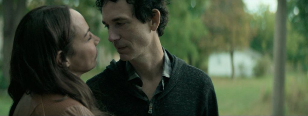 A still from Ashgrove. Jennifer and Jason stand close to each other and look in each other's eyes as they stand among trees. 
