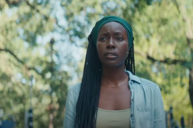 A still from Nanny. Aisha stands among trees with a concerned look on her face. She wears a dark green headwrap around her long black braids. She has a light green tank top and a light blue button down shirt open on top of it. 
