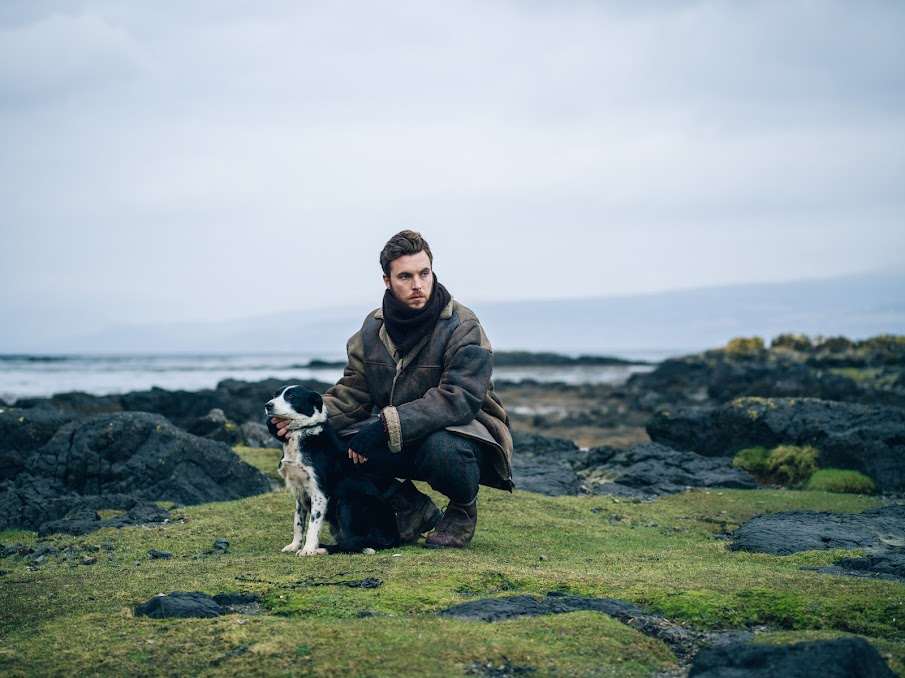 A still from Shepherd. Eric kneels on a patch of rocky grass holding his dog. The sea and a grey horizon are visible behind him. 