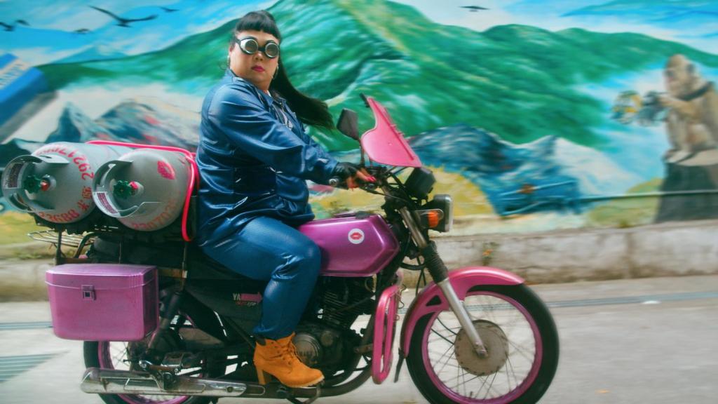 A still from Tank Fairy. The Tank Fairy rides a pink motorbike and wears a shiny blue jumpsuit and high-heeled work boots. She drives past a wall mural depicting mountains. 