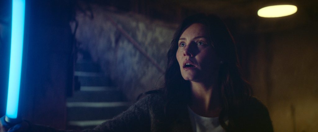 A still from The Cellar. Keira holds a blacklight that glows blue as she stands in her shadowy cellar. She gazes in fear at what the blacklight reveals. 
