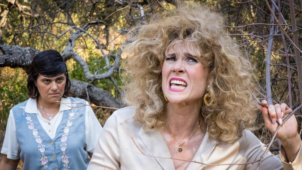 A still from Wild Bitch. Barb stands behind Melanie, watching her intently as she grimaces with her mouth open. They are walking through a heavily wooded area. 