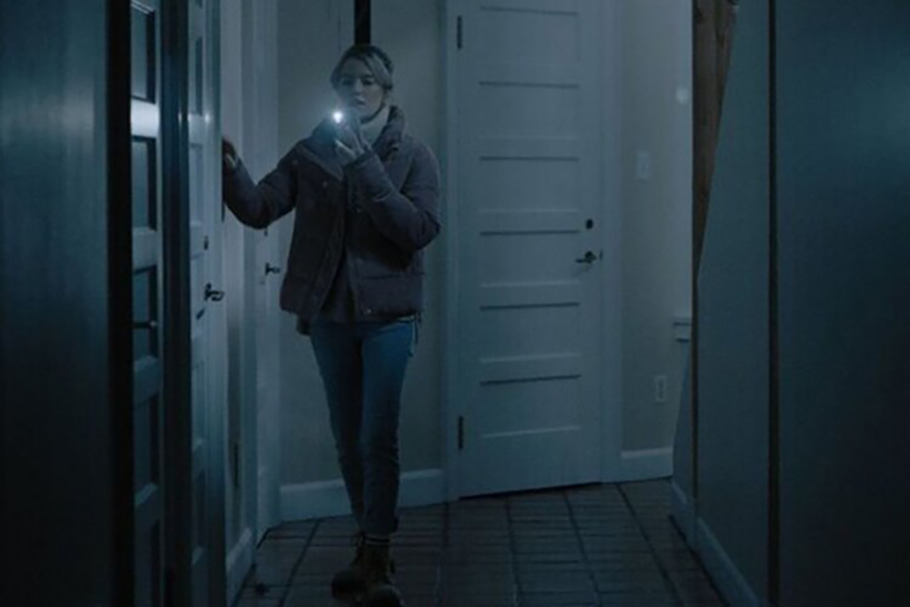 A still from See for Me. Sophie walks down a hallway, running her hand along the wall and using her other hand to hold up a cell phone with its flashlight on.