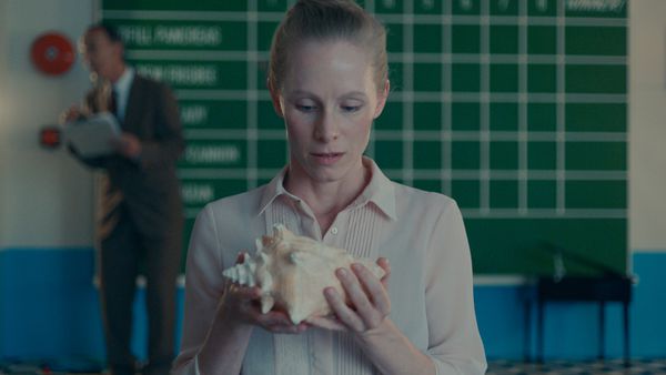 A still from Stanleyville. Maria stares at a conch shell in her hands while Homunculus stands behind her in front of a scoreboard.