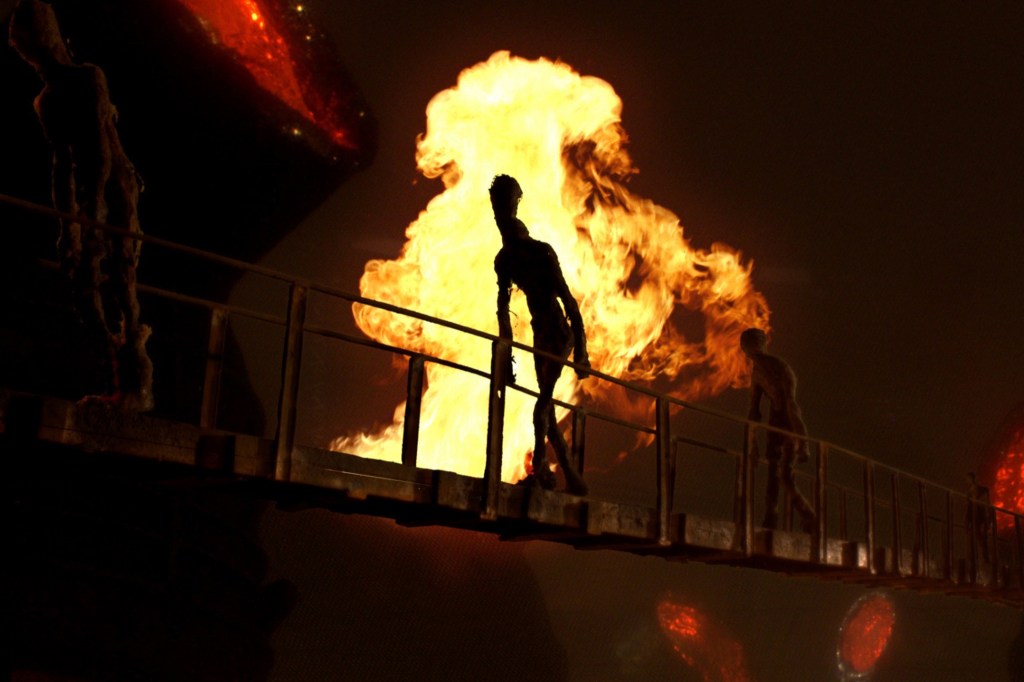 A still from Mad God. Elongated, human-like figures trudge across a drawbridge as flames erupt behind them. 