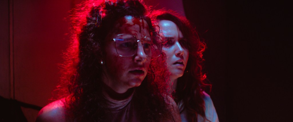 A still from Revealer. Sally and Angie stand in a red-lit room looking fearfully ahead as blood runs down their foreheads. 