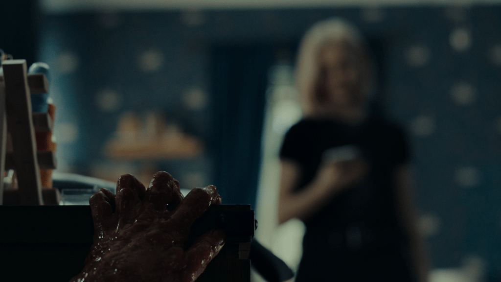A still from 'Appendage.' A slimy hand grasps a desk in the foreground as Hannah checks her phone in the background. 