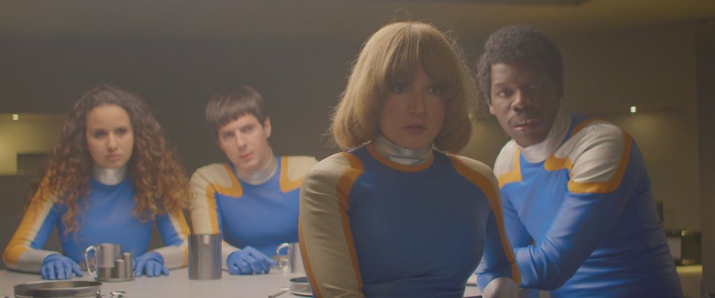 A still from Smoking Causes Coughing. Four superheroes in blue spandex suits sit at a table looking at someone off-camera in surprise. 